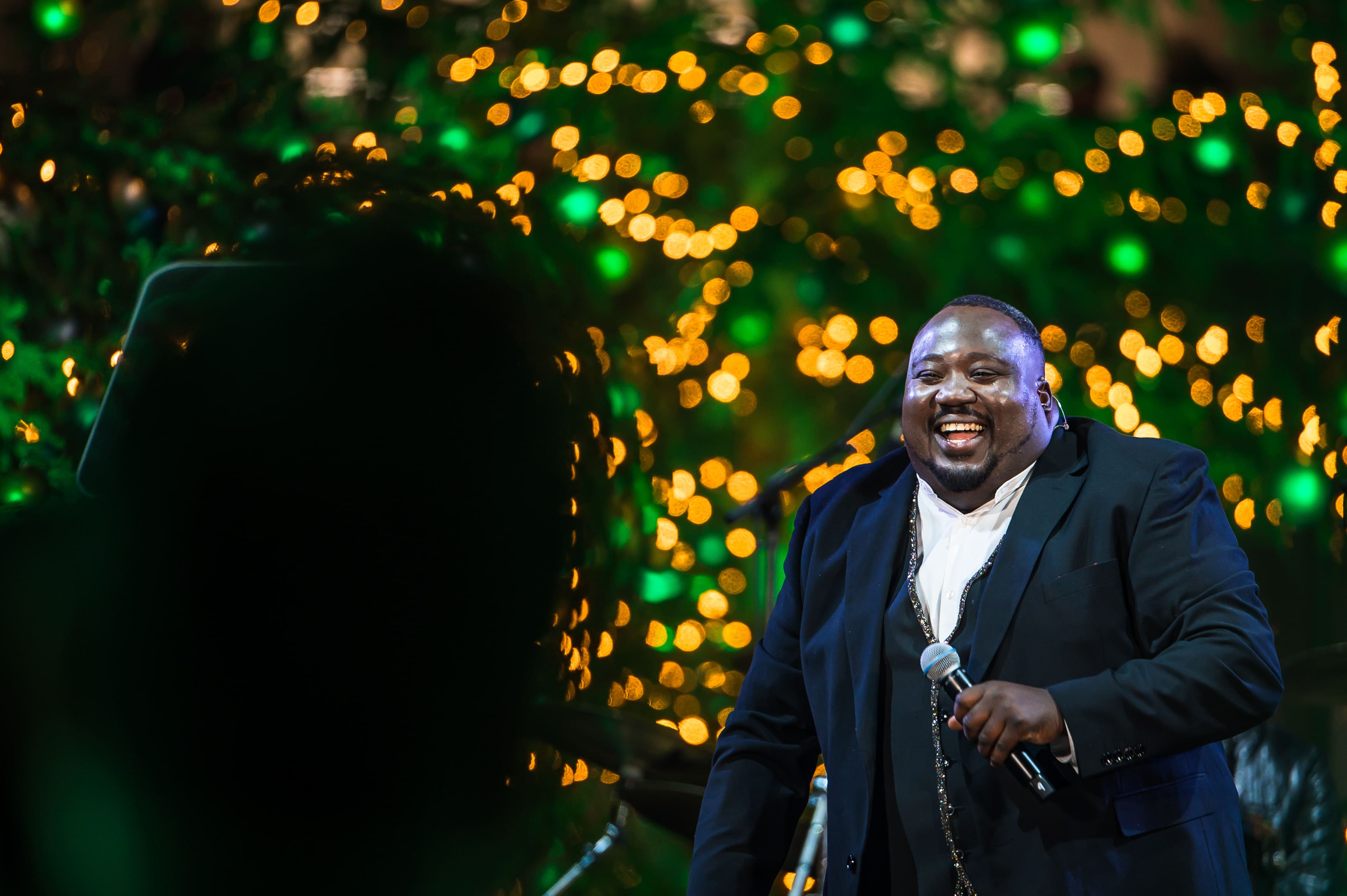 Ο παρουσιαστής της αντίστροφης μέτρησης, που χαμογελάει μπροστά από φωτισμένα χριστουγεννιάτικα δέντρα κρατώντας το μικρόφωνο.