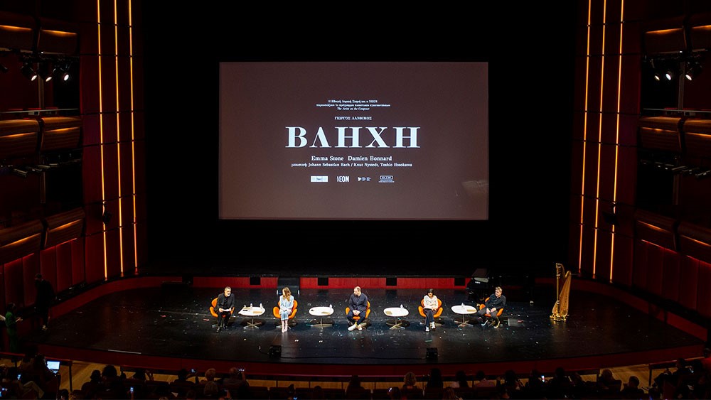 Φωτογραφία από τη κεντρική σκηνή της Εθνικής Λυρικής Σκηνής στην οποία κάθονται πέντε άτομα και συζητάνε για το έργο ΒΛΗΧΗ όπως φαίνεται και στην οθόνη που προβάλεται από πίσω τους 