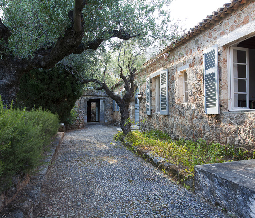 Φωτογραφία που δείχνει ένα πέτρινο διάδρομο να βρίσκεται ανάμεσα από ένα πέτρινο σπίτι και από τα δέντρα και το πράσινο