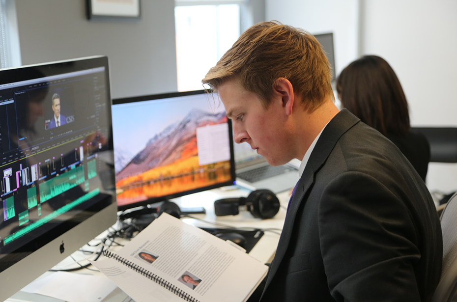 Ένας νεαρός άνδρας με κοστούμι κάθεται μπροστά από έναν υπολογιστή με λογισμικό επεξεργασίας και διαβάζει ένα εγχειρίδιο