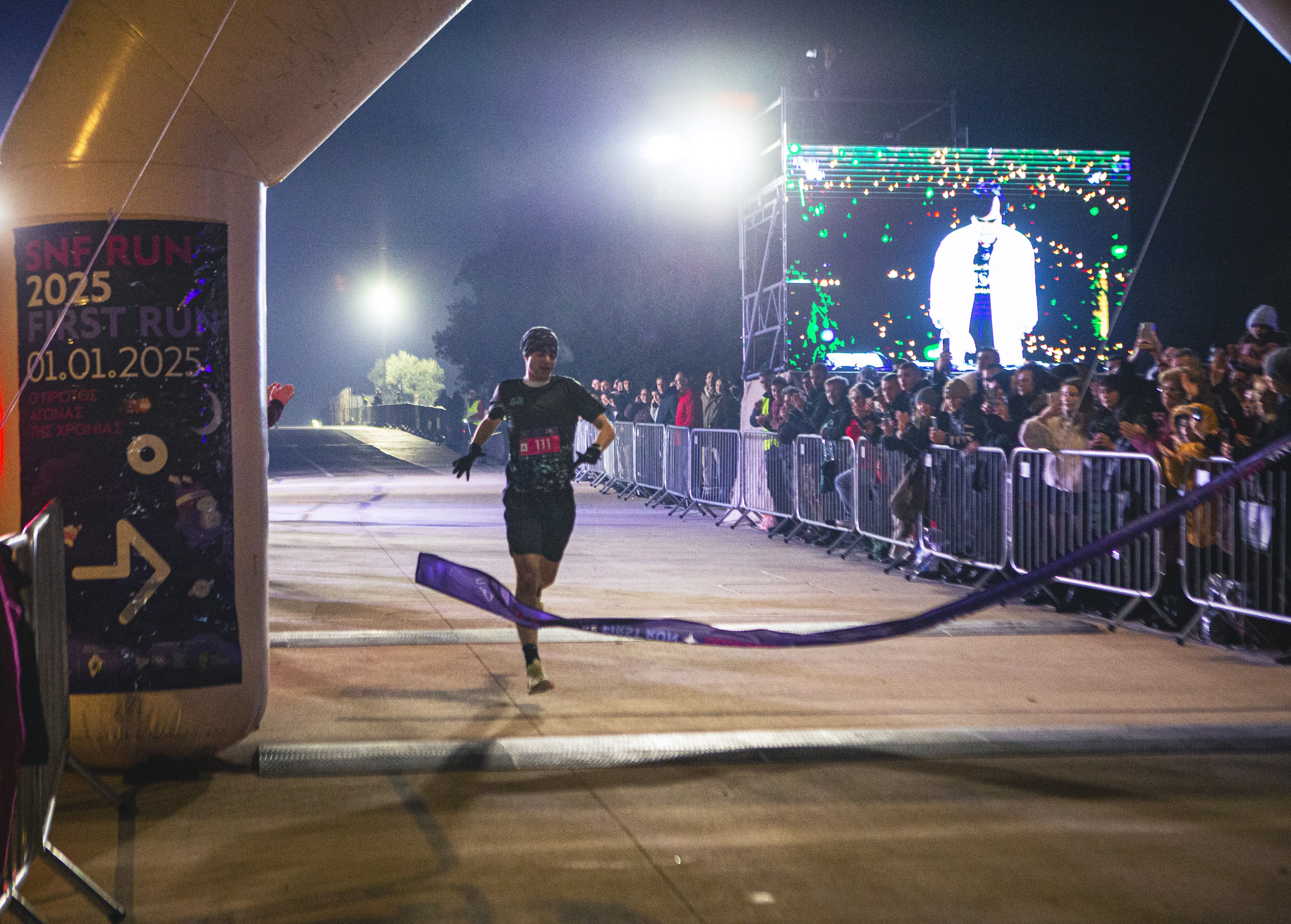 Ο πρώτος δρομέας του SNF First Run διασχίζοντας τη γραμμή τερματισμού, φωτισμένος από δυνατούς προβολείς.