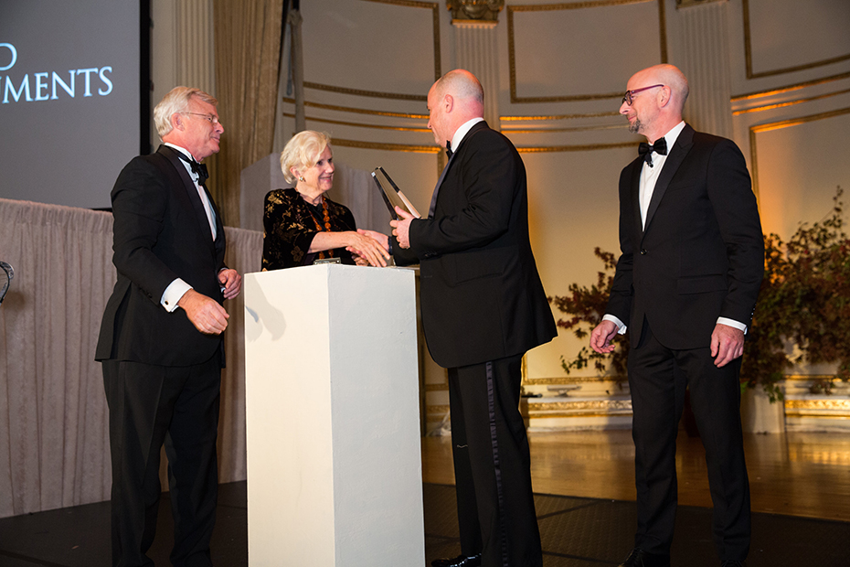 Ο Αντρέας Δρακόπουλος, Πρόεδρος του Ιδρύματος Σταύρος Νιάρχος, κρατάει ένα βραβείο τιμής και κάνει μια χειραψία με μια γυναίκα και δύο άντρες δίπλα τους τους παρακολουθούν