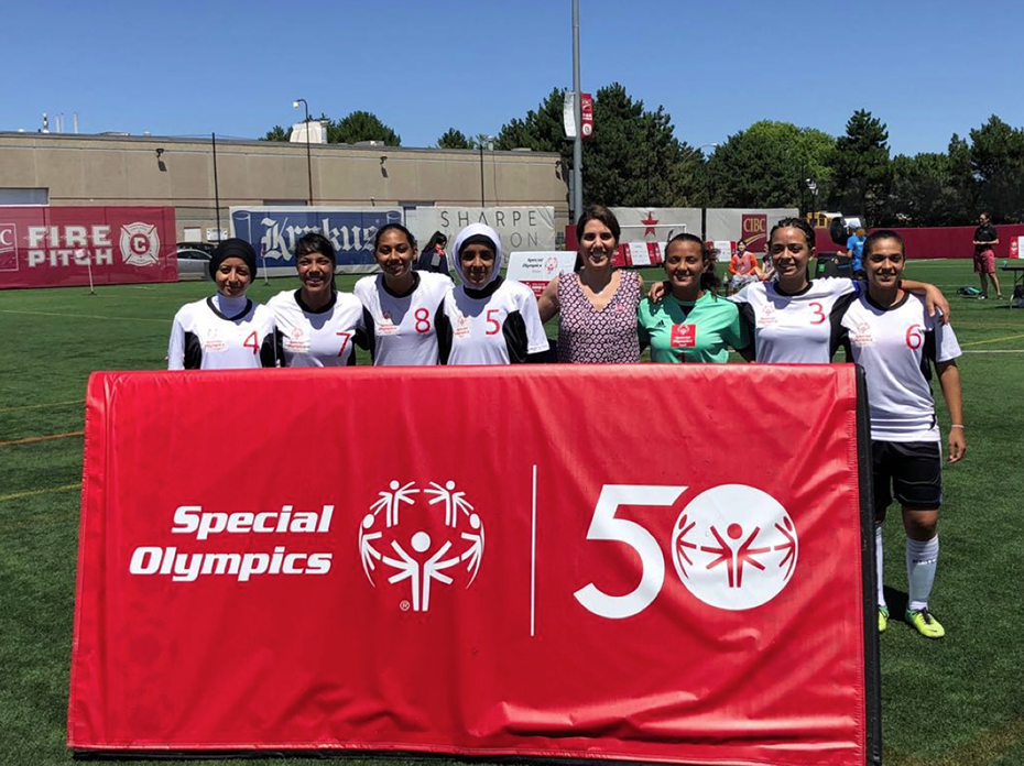 A team of young women wearing team jerseys stand together behind a Special Olympics banner in a soccer field