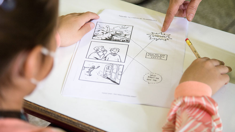 Ένα μικρό κοριτσάκι κρατάει ένα μολύβι και ένα χέρι της δείχνει κάτι στο χαρτί που έχει μπροστά της 