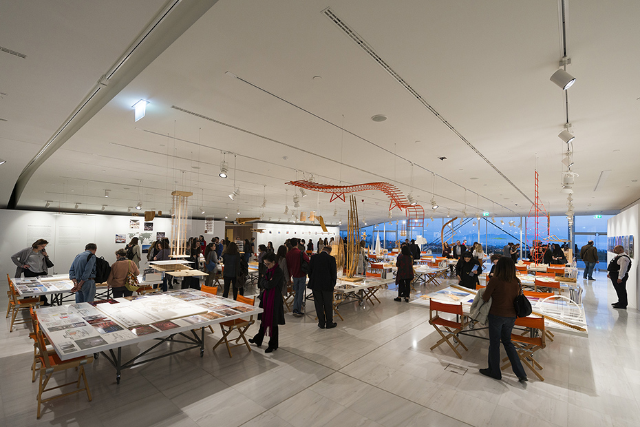 Μακρινη Φωτογραφία που δείχνει κόσμο να στέκεται και να κοιτάει τραπέζια στα οποία υπάρχουν σχέδια, υπομνήματα, εικόνες και μακέτες κτηρίων 