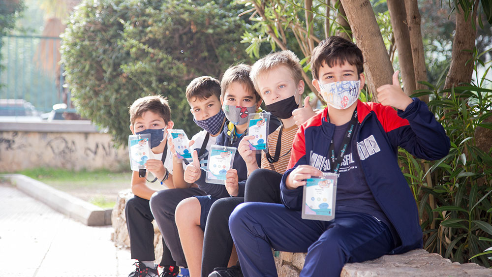 Πέντε μικρά παιδιά που φοράνε μάσκα κάθονται σε ένα πεζούλι και κρατάνε το καρτελάκι τους ποζάροντας για τη φωτογραφία  