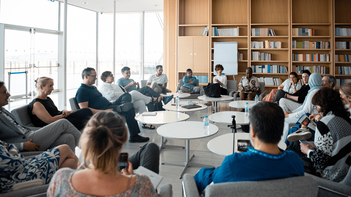 Δεκαοκτώ άτομα κάθονται σε έναν μεγάλο κύκλο σε έναν φωτεινό χώρο με βιβλιοθήκες που βρίσκονται σε ένα μέρος του τοίχου στο βάθος.