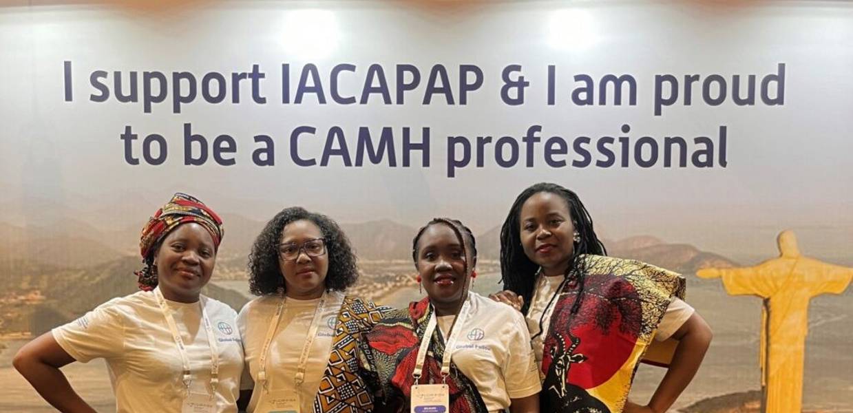 Four women smile for the camera wearing traditional African clothing with colorful motifs, in front of a banner supporting an organization.