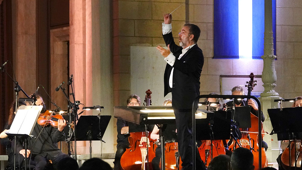 Ένας μαέστρος στο κέντρο της σκηνής κατευθύνει τα μέλη της ορχήστρας που κάθονται στις θέσεις τους με τα μουσικά τους όργανα 