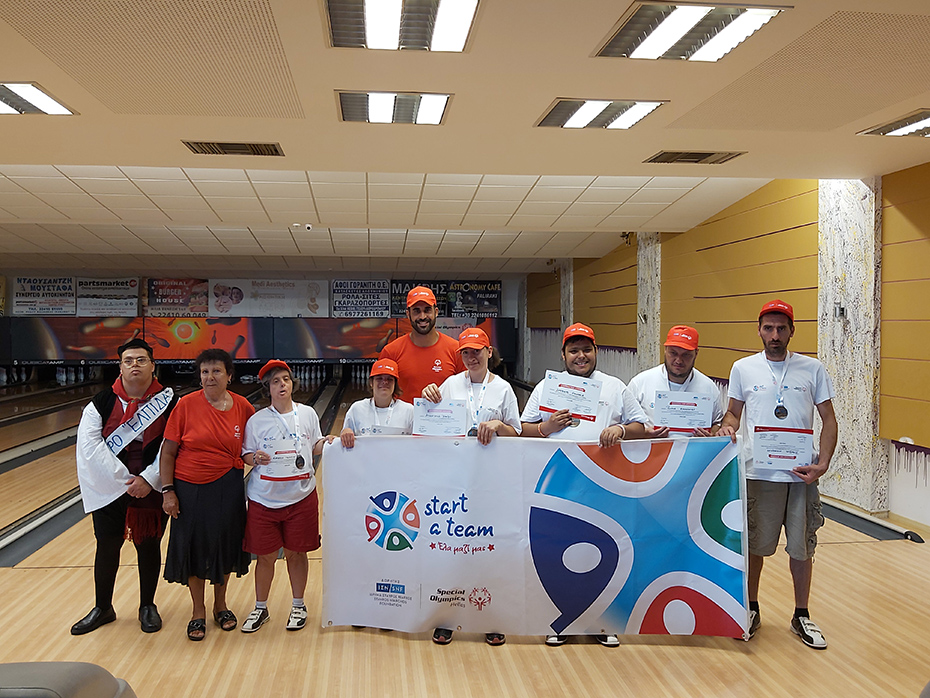 Μια ομάδα ανθρώπων στέκονται μπροστά από έναν διάδρομο bowling και κρατάνε ένα πανό το οποίο γράφει start a team, έλα μαζί μας