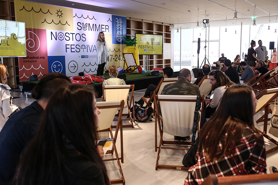 Μια γυναίκα δίνει ομιλία στο Summer Nostos Festival, ενώ το κοινό κάθεται στις καρέκλες του γκαζόν 