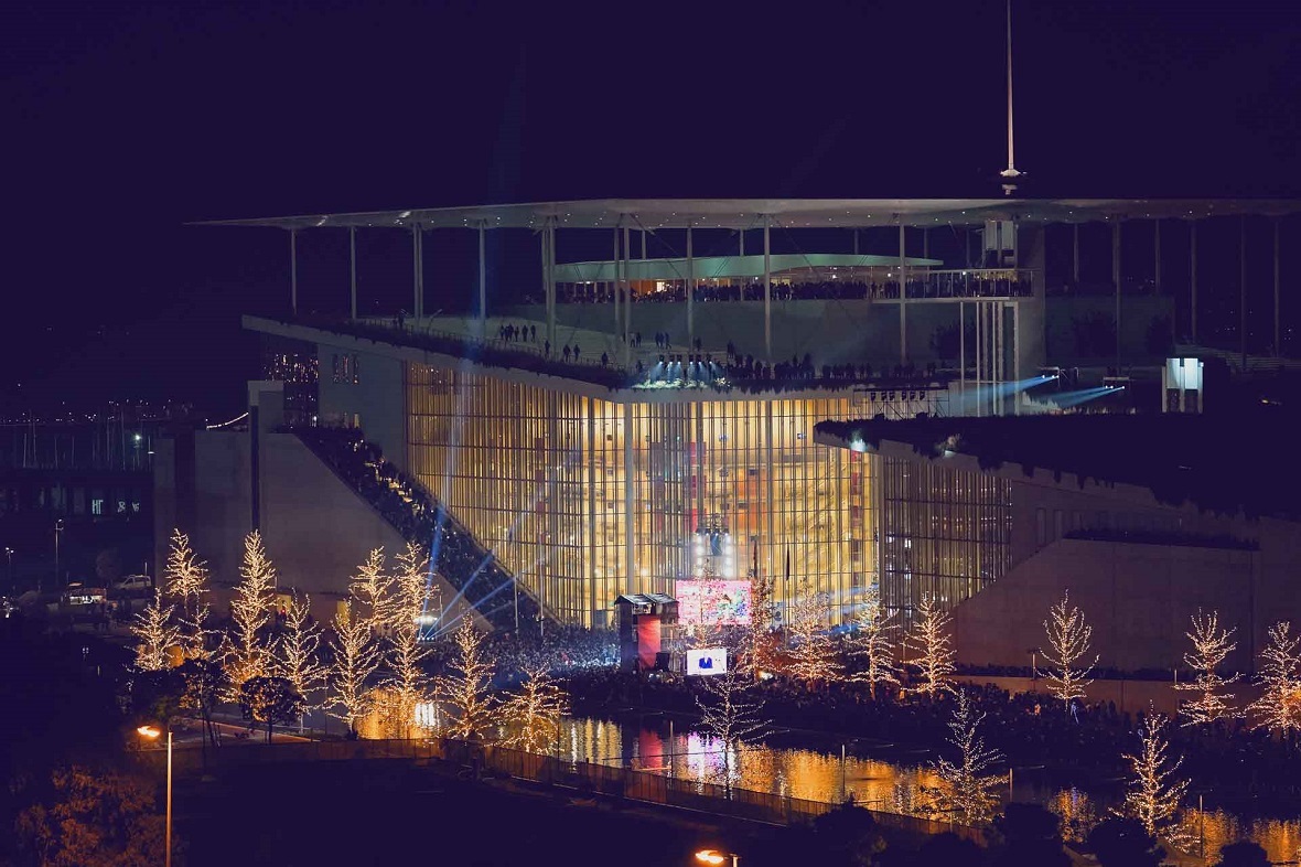 Φωτογραφία τραβηγμένη το βράδυ η οποία δείχνει χριστουγεννιάτικα στολισμένα δέντρα γύρω από μια λίμνη και ένα μεγάλο γυάλινο κτήριο γεμάτο κόσμο 