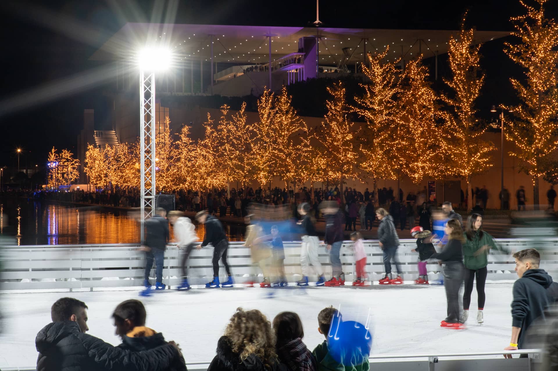 Άνθρωποι στο παγοδρόμιο του Κέντρου Πολιτισμού Ίδρυμα Σταύρος Νιάρχος το οποίο είναι τοποθετημένο μπροστά από το Κανάλι και πολλά δέντρα γύρω του να είναι στολισμένα με χριστουγεννιάτικα λαμπάκια.