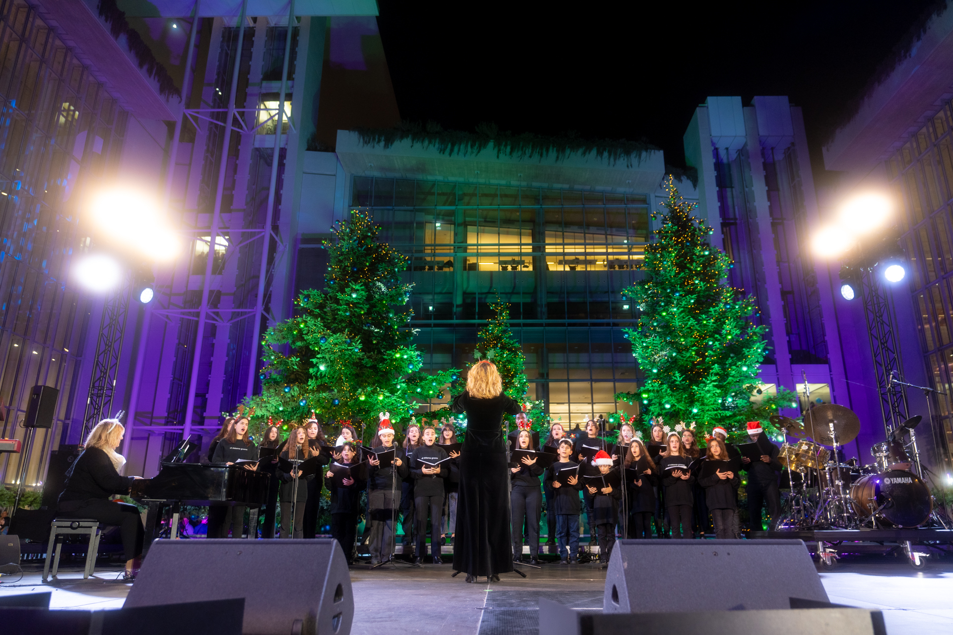 Στη σκηνή μια χορωδία παιδιών τραγουδούν ακολουθώντας τις οδηγίες της μαέστρου και της πιανίστριας και φορούν χριστουγεννιάτικους σκούφους, ενώ πίσω τους υπάρχουν στολισμένα χριστουγεννιάτικα δέντρα. 