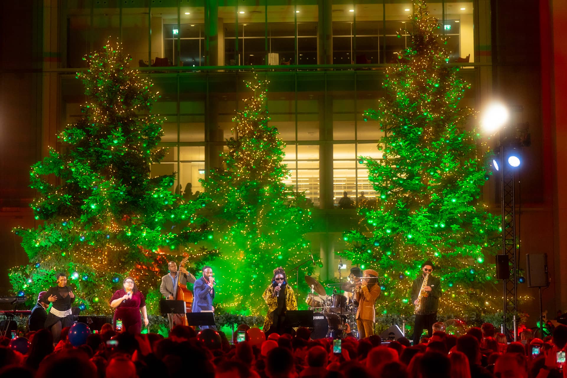 Μπάντα που παίζει μουσική και τραγουδά στη σκηνή, με φόντο στολισμένα χριστουγεννιάτικα δέντρα.