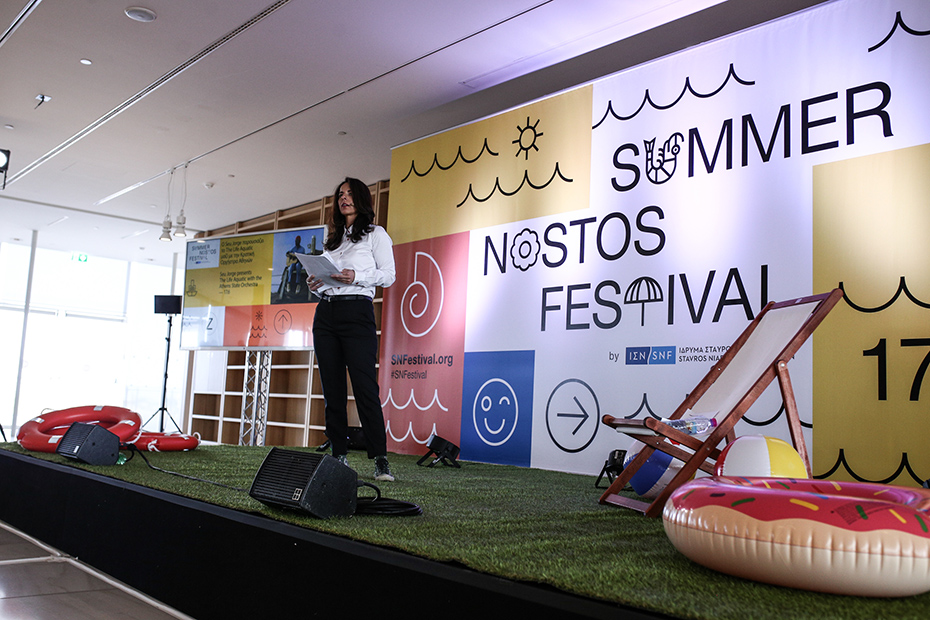 Μια γυναίκα μιλάει στη σκηνή του Summer Nostos Festival, η οποία διαθέτει τεχνητό γρασίδι, μπάλες θαλάσσης, κρίκους σωσιβίων και μια καρέκλα γκαζόν.