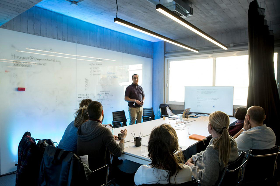 Σε μία αίθουσα κάθεται μια ομάδα ανθρώπων και παρακολουθούν έναν άντρα που τους μιλάει
