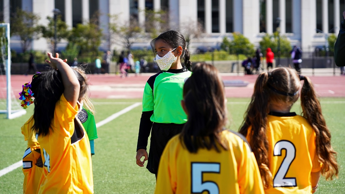Μικρά κορίτσια που φοράνε κίτρινη ποδοσφαιρική στολή είναι μαζεμένες και ακούν την κοπέλα που έχει τον ρόλο του διατητή φορώντας πράσινη ποδοσφαιρική στολή