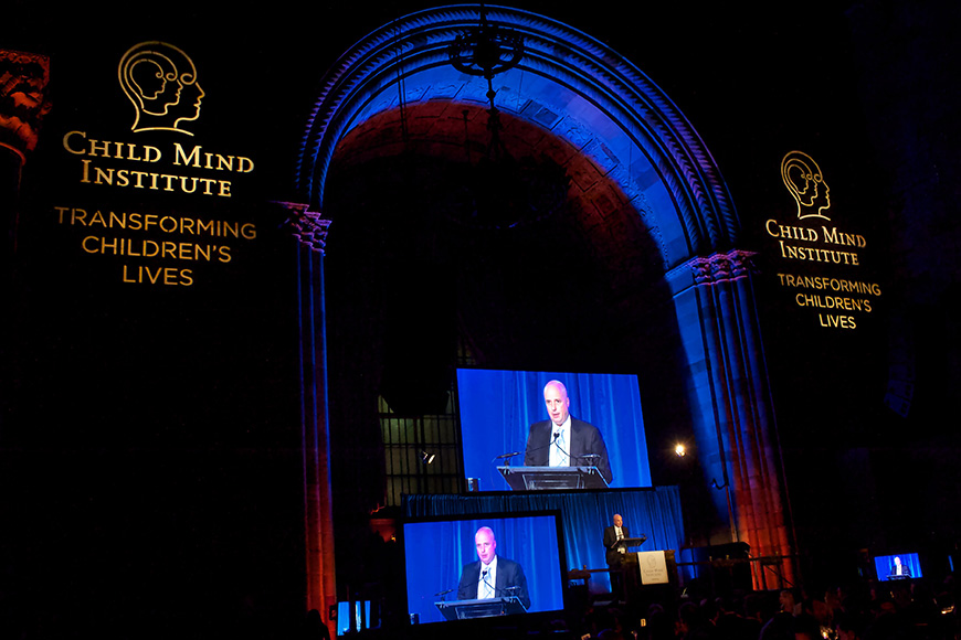 Μακρινή φωτογραφία από τη σκηνή η οποία μοιάζει σαν πύλη στην οποία ο Αντρέας Δρακόπουλος, Πρόεδρος του Ιδρύματος Σταύρος Νιάρχος βγάζει λόγο και δίπλα του υπάρχουν οθόνες που προβάλλουν την ομιλία του ενώ η σκηνή γράφει CHILD MIND INSTITUTE TRANSFORMING CHILDREN'S LIVES 