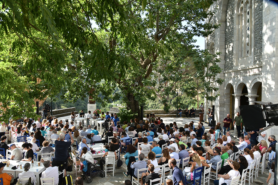 Μακρινή φωτογραφία που δείχνει ένα πλήθος ανθρώπων να κάθονται στην κεντρική πλατεία του χωριού και να παρακολουθούν μια συζήτηση