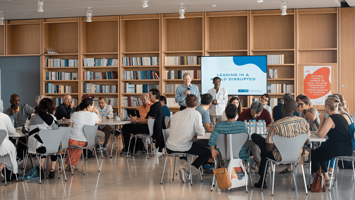 Άνθρωποι καθισμένοι σε τραπέζια σε μια βιβλιοθήκη, περιτριγυρισμένοι από ράφια γεμάτα βιβλία.