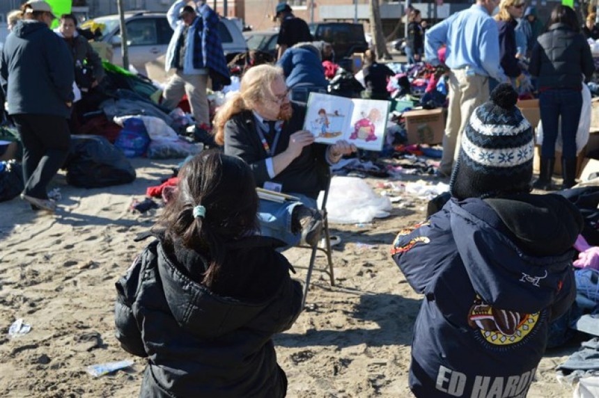 Ένας άντρας κάθεται και δείχνει εικόνες από ένα βιβλίο σε μικρά παιδιά που μένουν στον δρόμο με τις οικογενειές τους