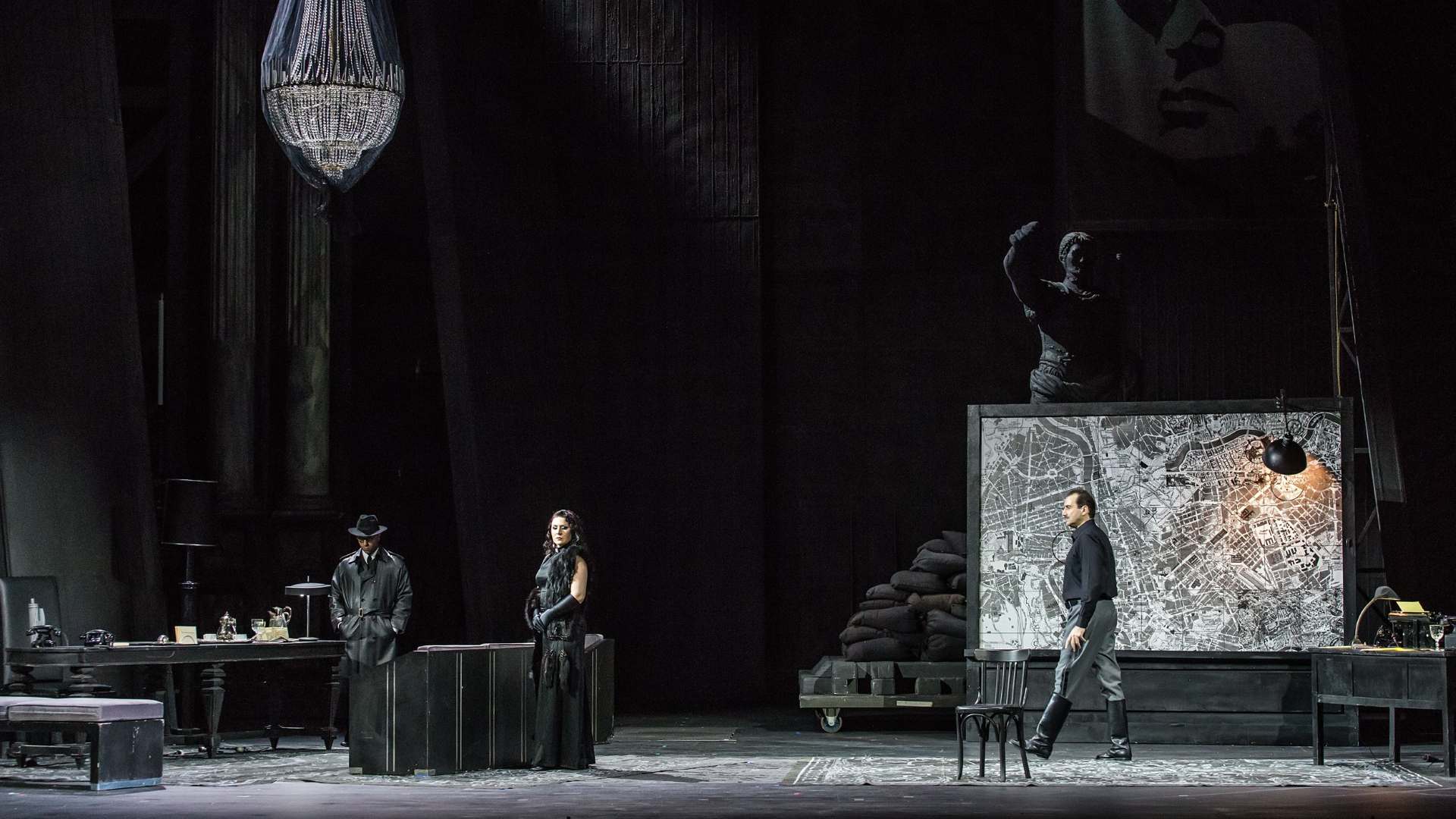 A dark stage setting with three actors: two men—one in motion, the other still, dressed in black clothes and hat—and a woman with a mysterious presence.