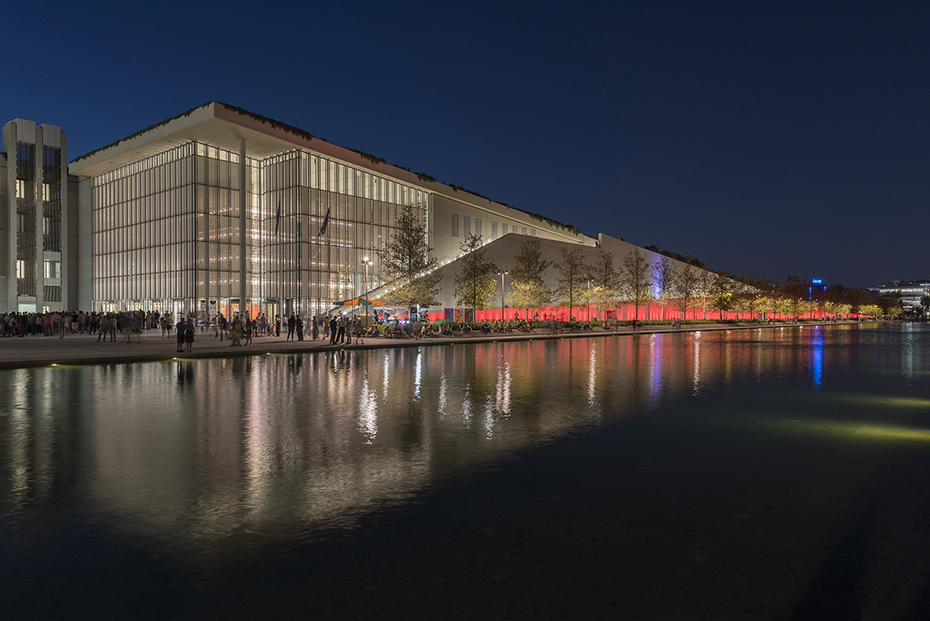 Φωτογραφία τραβηγμένη τη νύχτα η οποία δείχνει πλήθος ανθρώπων να περπατάει στο πεζοδρόμιο ανάμεσα σε μια λίμνη και ένα μεγάλο γυάλινο κτήριο 