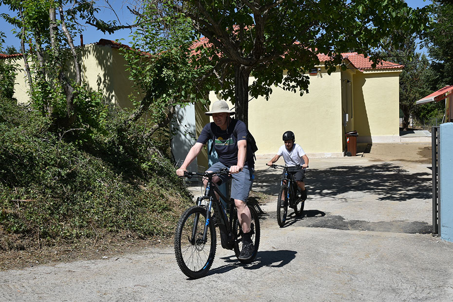 Ένας άντρας φοράει καπέλο και κάνει ποδήλατο ενώ ένα παιδάκι τον ακολουθεί με το δικό του ποδήλατο 