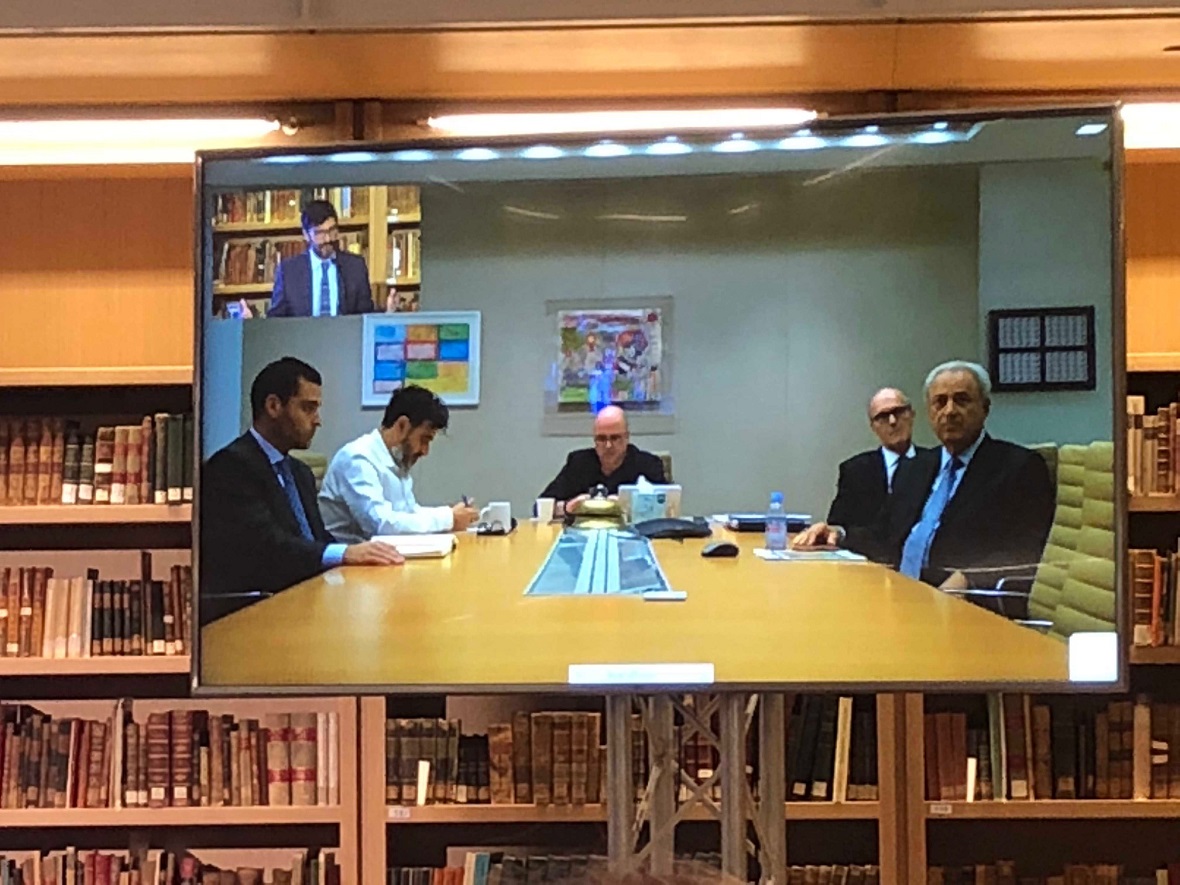 Φωτογραφία που δείχνει την οθόνη στην οποία προβάλλεται μια ζωντανή σύνδεση. Από τη μια πλευρά φαίνονται πέντε άντρες καθισμένοι γύρω από ένα τραπέζι συνεδριάσεων και από την άλλη φαίνεται ένας άντρας που μιλάει 