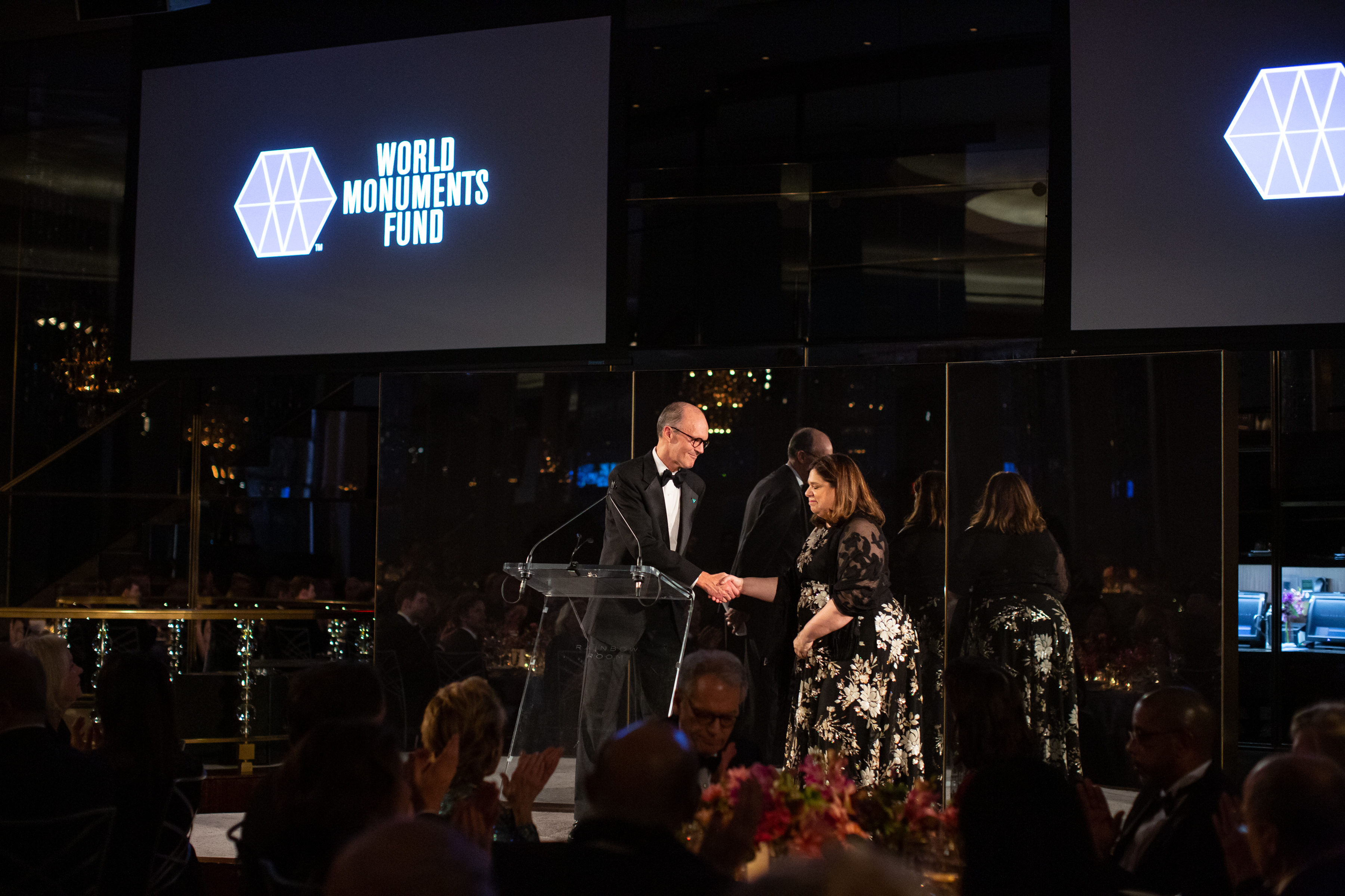 Ένας άνδρας σφίγγει το χέρι μιας γυναίκας στη σκηνή στο Hadrian Gala 2018 του Παγκόσμιου Ταμείου Μνημείων