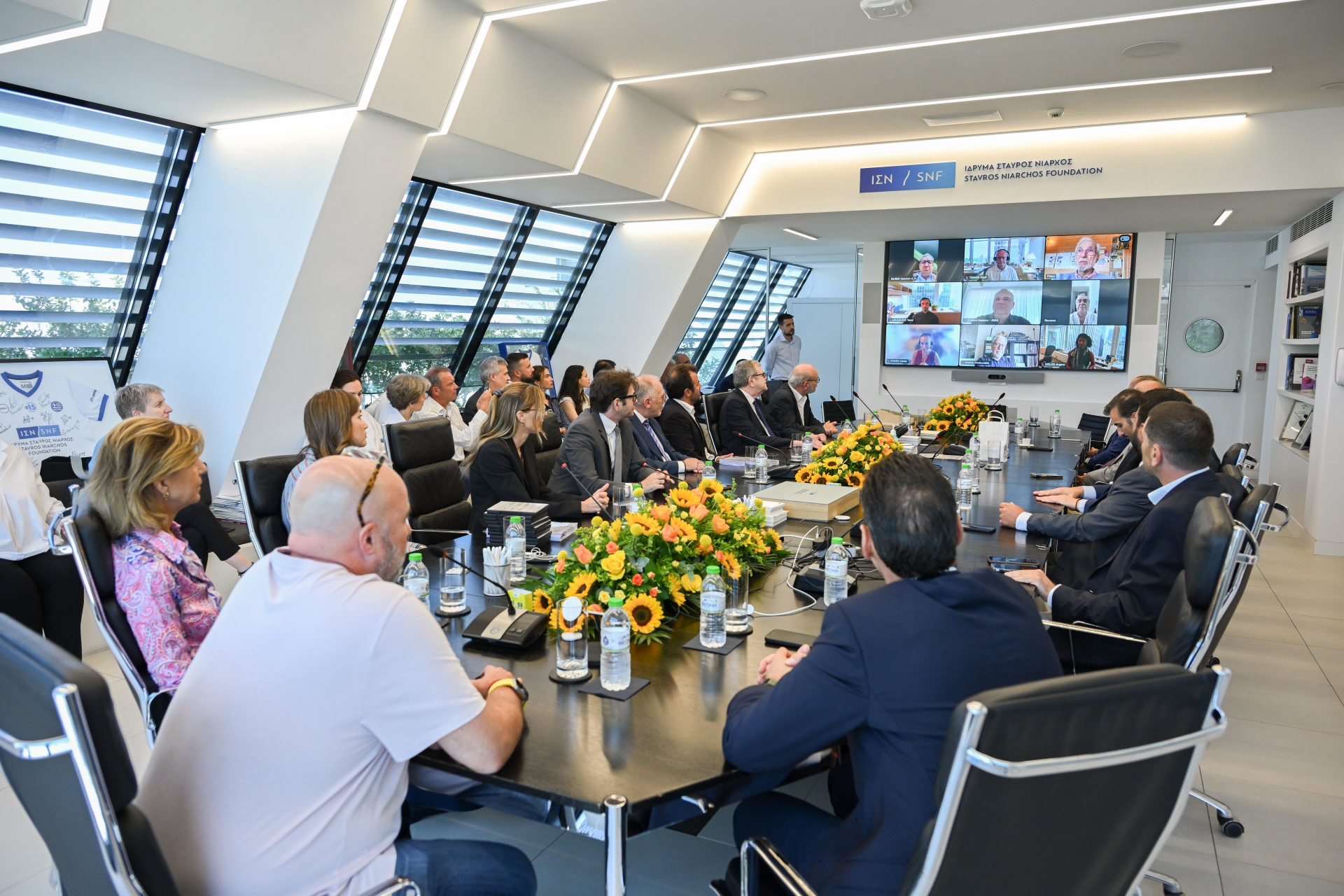 Πολλά μέλη του Ιδρλυματος Σταύρος Νιάρχος κάθονται γύρω από ένα τραπέζι και συζητούν με άλλα μέλη του Ιδρύματος μέσω διαδικτυακής σύνδεσης 