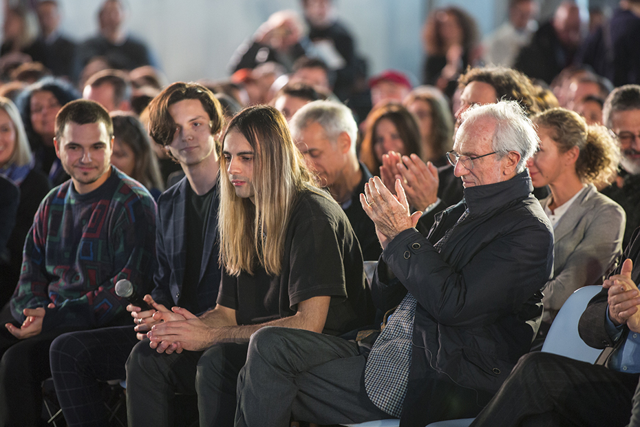 Κοντινή φωτογραφία από το κοινό η οποία δείχνει έναν νεαρό άντρα να κρατάει ένα μικρόφωνο και οι υπόλοιποι να χειροκροτούν
