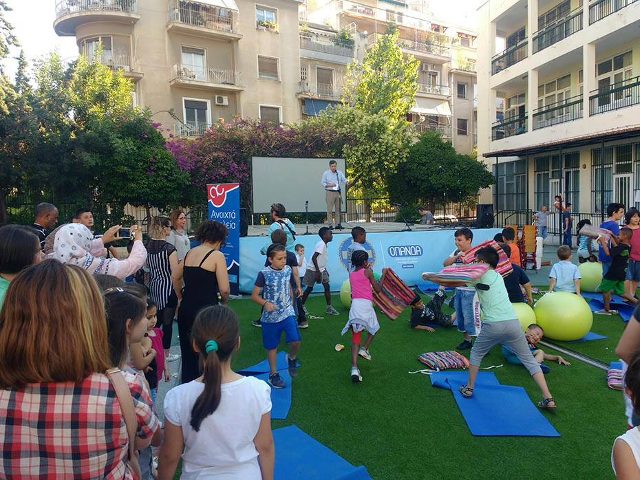 Στον προαύλιο χώρο ενός σχολείου έχει στηθεί μια εξέδρα και γρασίδι και τα παιδιά παίζουν ενώ κόσμος τους παρακολουθεί 