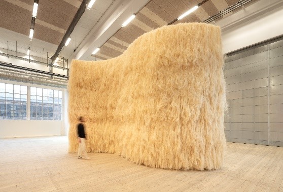 Artistic installation in a wave shape of imposing size, covered on the outside with material in the texture and color of straw, while a man in motion seems to pass by.