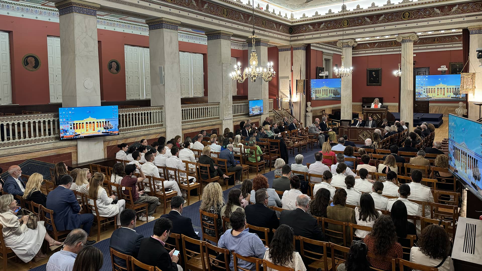 Πολλά άτομα καθισμένα σε μια ευρύχωρη αίθουσα παρακολουθούν μια ομιλία.