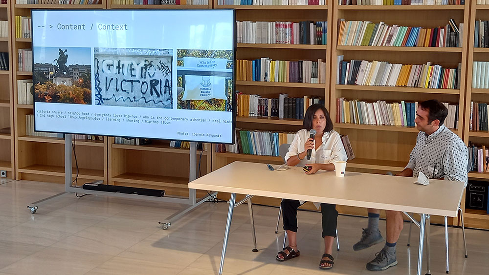 Ένας άντρας και μια γυναίκα κάθονται σε ένα τραπέζι και μιλούν έχοντας δίπλα τους μια οθόνη να προβάλει το περιεχόμενο της συζήτησης 