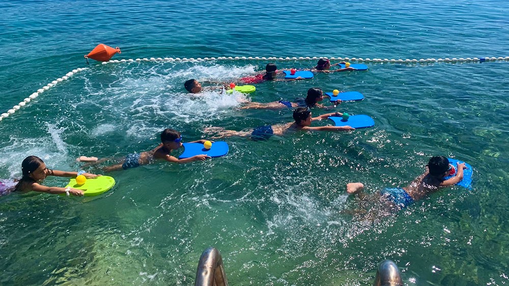 Μια ομάδα παιδιών μαθαίνουν να κολυμπούν στη θάλασσα κρατώντας σανίδες 