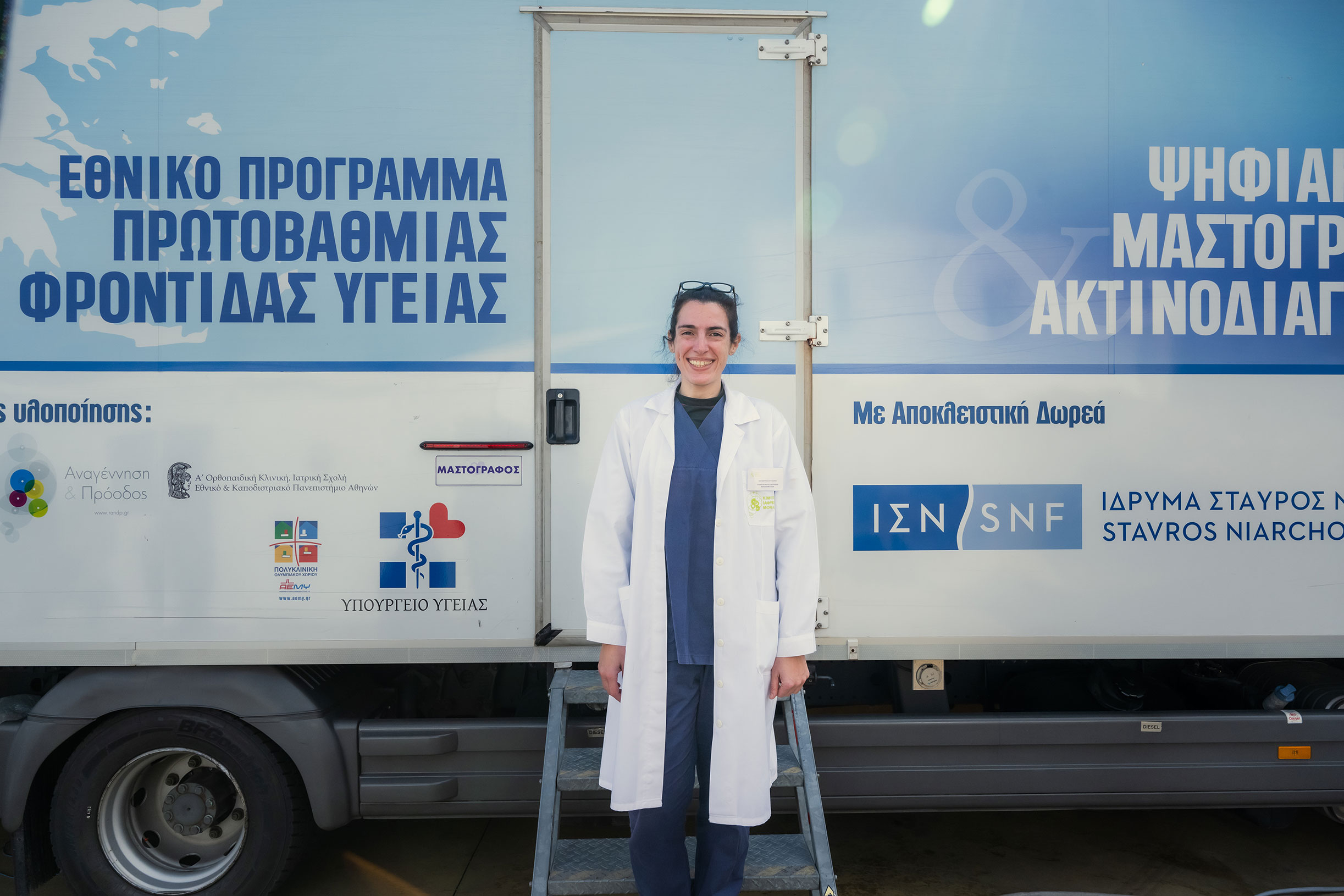 Μια γιατρός στέκεται μπροστά από την Κινητή Ιατρική Μονάδα και χαμογελάει.