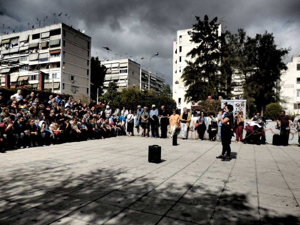 Μια γυναίκα μιλάει μπροστά σε ένα καθισμένο κοινό σε πλατεία γειτονιάς