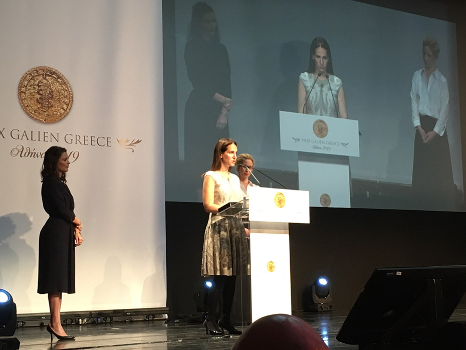 Τρεις γυναίκες στη σκηνή, η μία μιλάει στο βήμα και οι άλλες δύο στέκονται δεξιά και αριστερά από το βήμα, ενώ η οθόνη προβάλει τις τρεις γυναίκες 