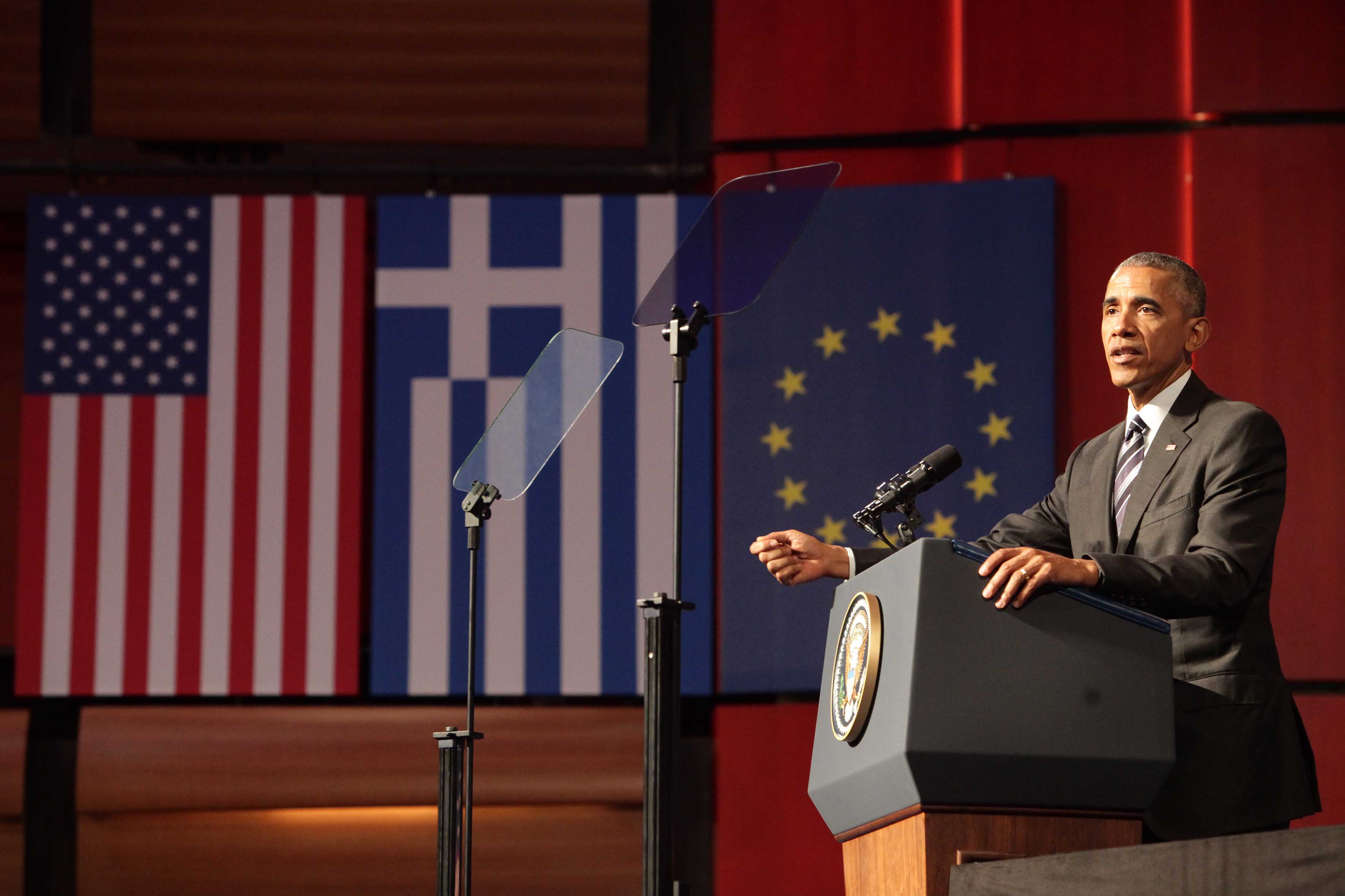 Ο πρόεδρος Ομπάμα μιλάει πίσω από ένα βήμα, ενώ στο βάθος διακρίνονται η σημαία των Ηνωμένων Πολιτειών, η ελληνική σημαία και η σημαία της Ευρωπαϊκής Ένωσης.