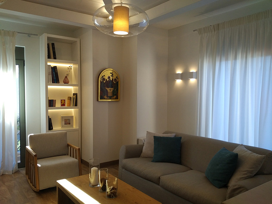 Μια φωτογραφία του Ξενώνα Ανακουφιστικής Φροντίδας, η οποία διαθέτει έναν καναπέ, βιβλιοθήκη και ζεστό φωτισμό