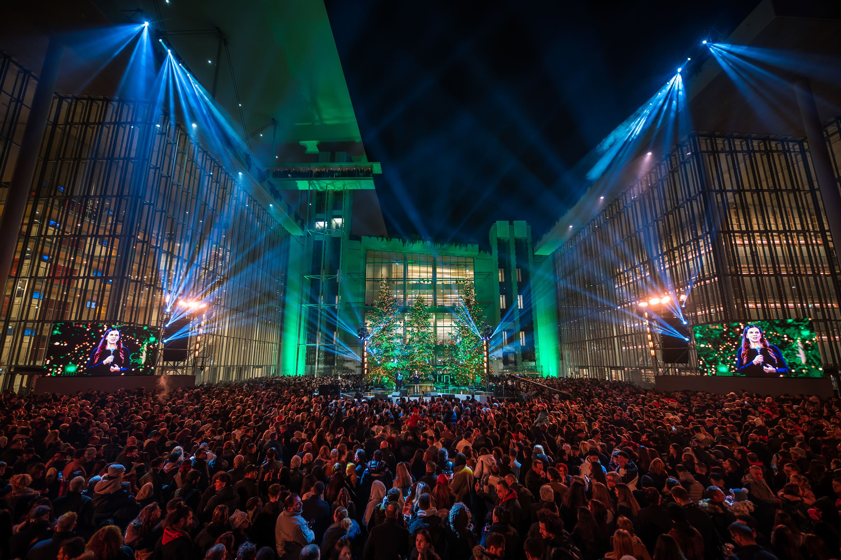 Η Αγορά του ΚΠΙΣΝ με δύο οθόνες που δείχνουν την παρουσιάστρια πάνω στη σκηνή, μπροστά από ένα μεγάλο πλήθος ατόμων.