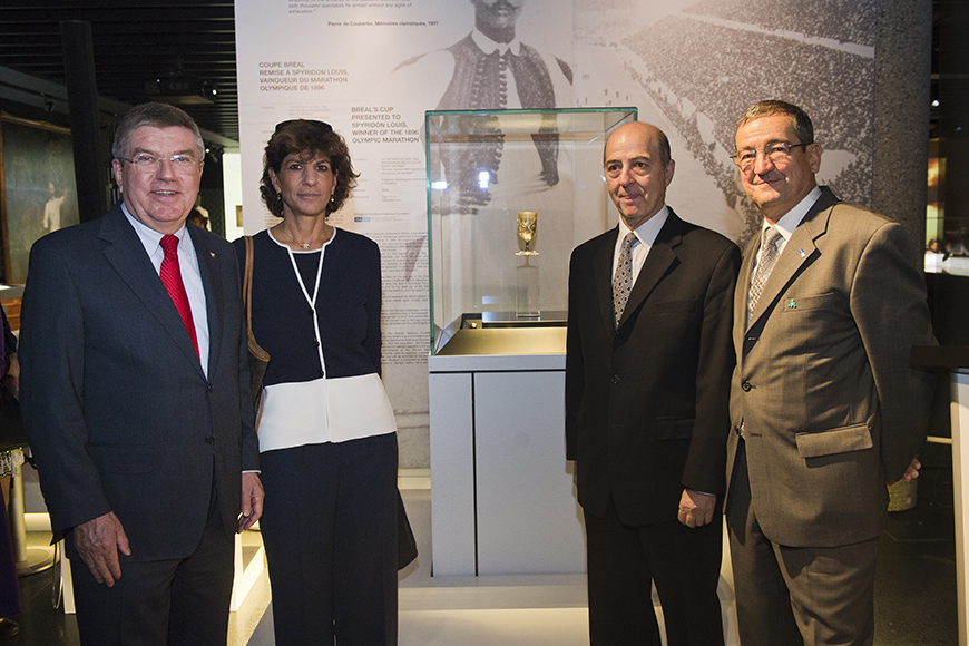 Τρεις άντρες και μια γυναίκα φωτογραφίζονται δίπλα από ένα ποτήρι παλάιότερης εποχής το οποίο είναι έκθεμα 