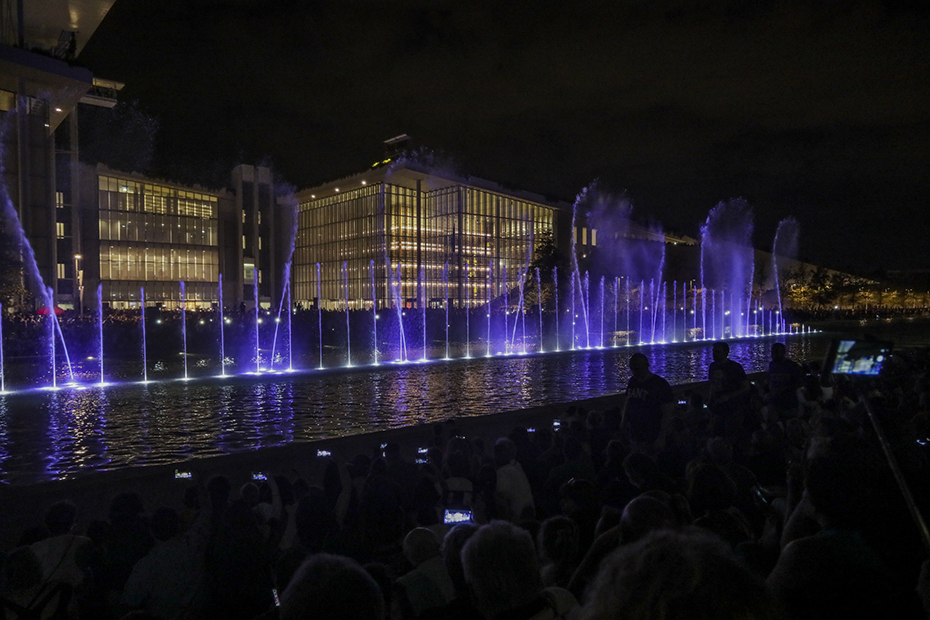 Κόσμος φωτογραφίζει το show που κάνει το συντριβάνι στο κέντρο μιας λίμνης μπροστά από ένα μεγάλο γυάλινο κτήριο