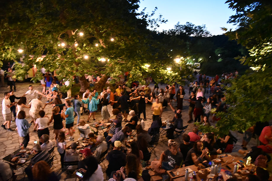 Φωτογραφία τραβηγμένη με drone η οποία δείχνει πλήθος ανθρώπων να κάθεται σε τραπέζια στην κεντρική πλατεία του χωριού, όσο άλλοι χορεύουν 