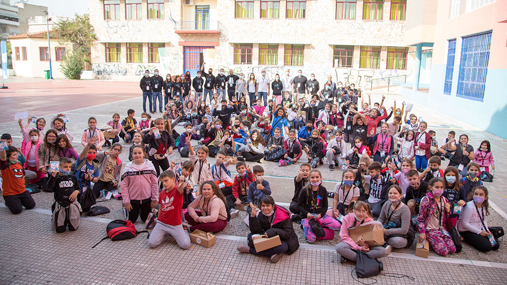 Πλήθος παιδιών κάθεται κάτω σε ένα γήπεδο και σηκώνουν τα χέρια τους στον αέρα για να βγάλουν μια αναμνηστική φωτογραφία 