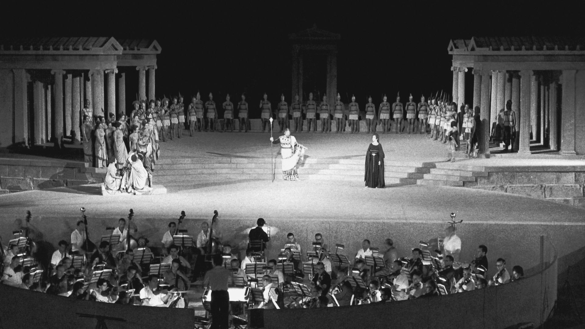 A theatrical scene on an ancient Greek-style stage with costumed actors and a live orchestra in front.
