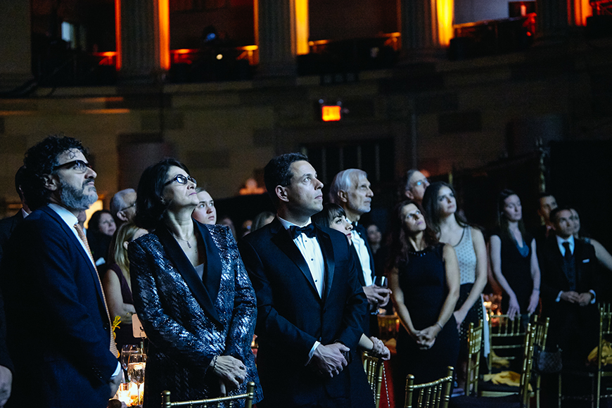 Φωτογραφία που δείχνει τον κόσμο να έχει σηκωθεί από τις καρέκλες τους και να παρακολουθεί κάτι το οποίο πραβάλλεται στην οθόνη